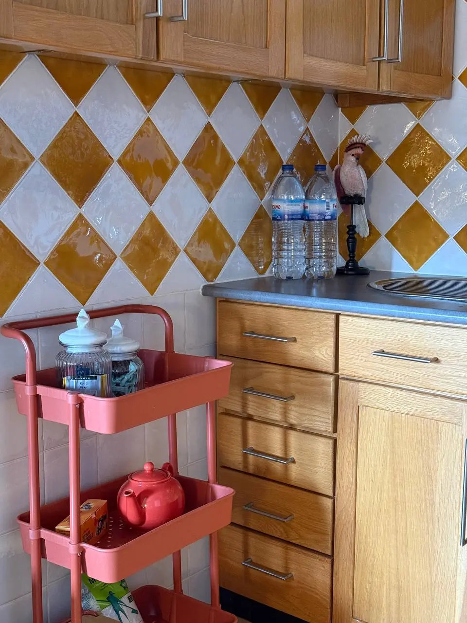 Kitchen counter detail with storage trolley, water bottles, drawers, and tiled wall